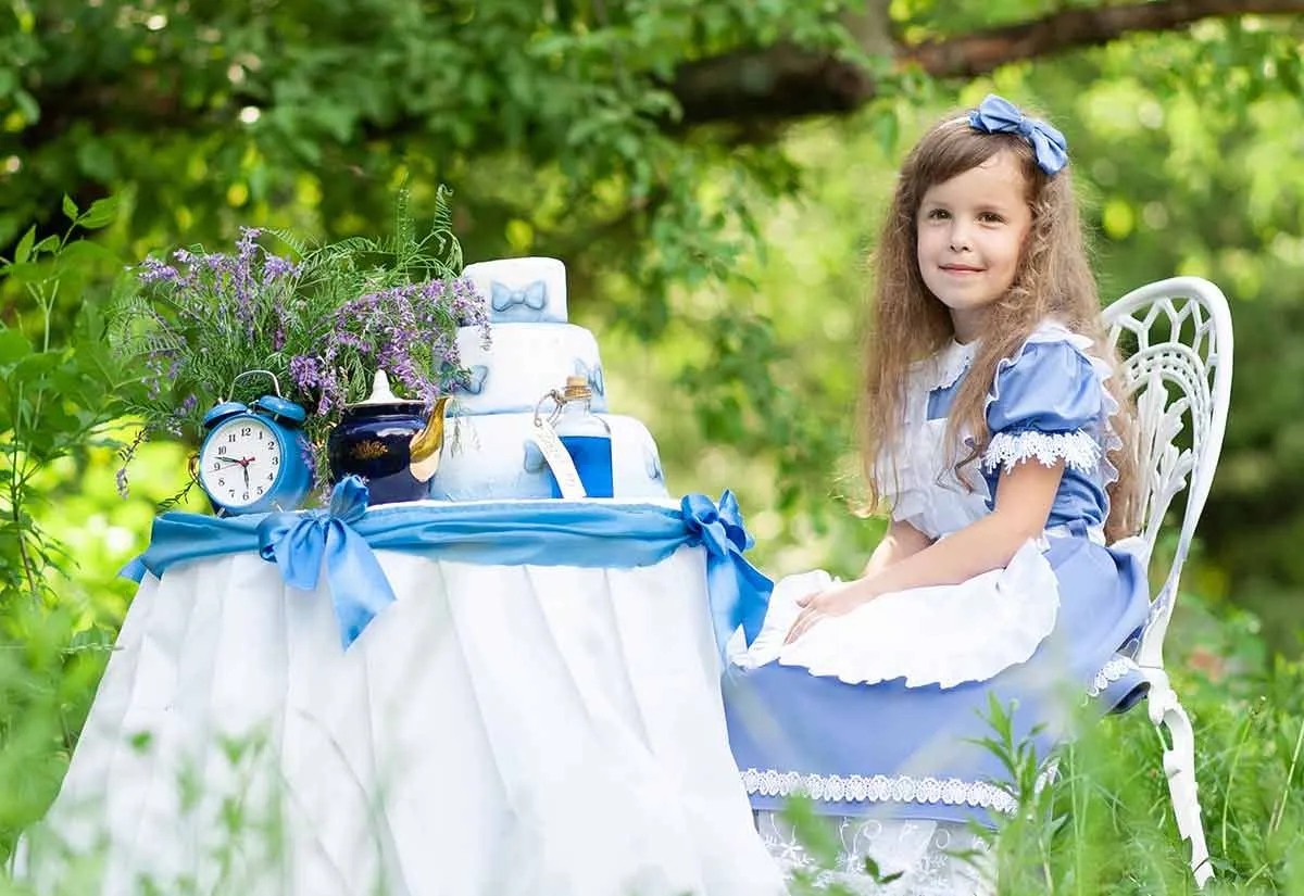 Маленька дівчинка, одягнена як Аліса в Країні Чудес, сиділа за столом на вулиці, готуючись до чаювання свого Божевільного Капелюшника.