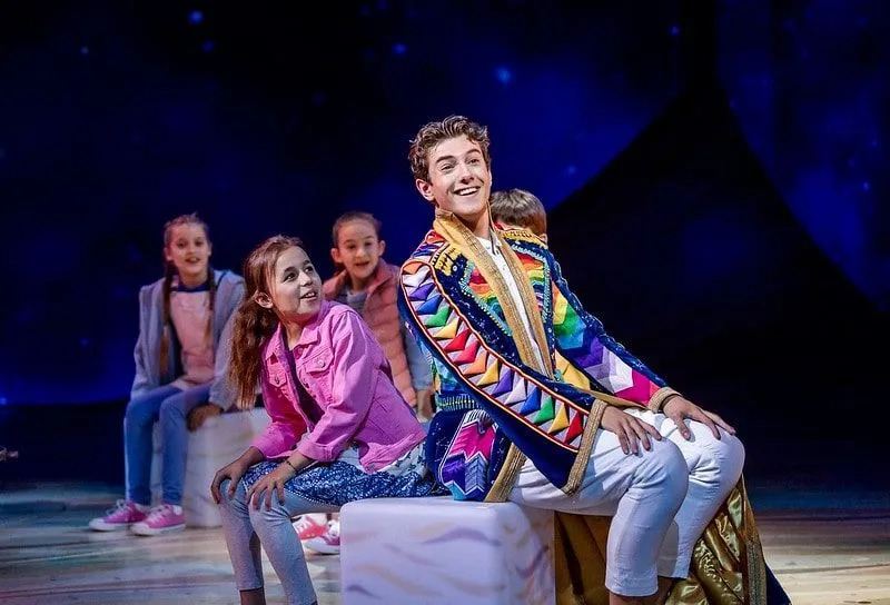 Die Hauptfigur Joseph in der Show Joseph and the Amazing Technicolor Dreamcoat trägt einen farbenfrohen Mantel und sitzt neben einer Gruppe von Kinderdarstellern. 