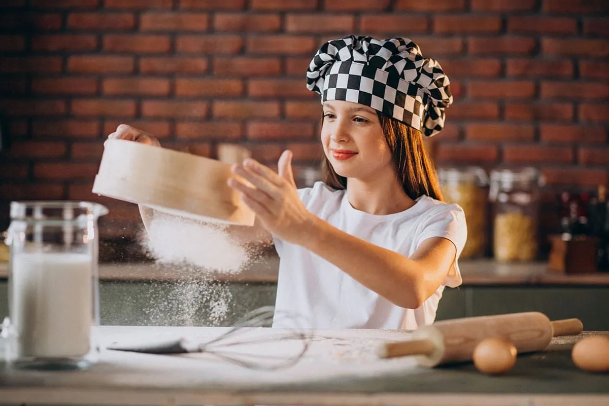 Mladá dívka prosévá mouku, aby udělala dort na pizzu, má na sobě černou a bílou kuchařskou čepici.