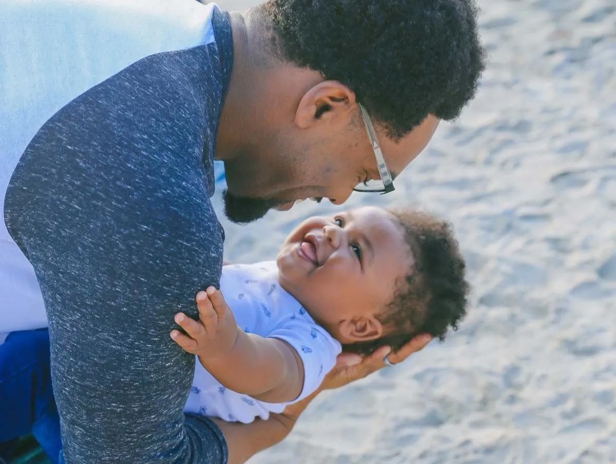 Táta na pláži drží dítě na zádech, zatímco se na něj usmívá, dítě mu úsměv opětuje.