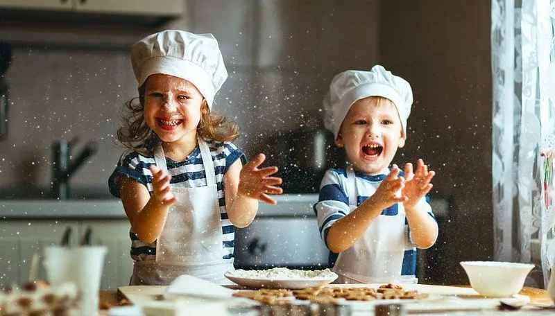Liten gutt og jente iført forkle og kokkehatter leker med mel mens de baker småkaker.