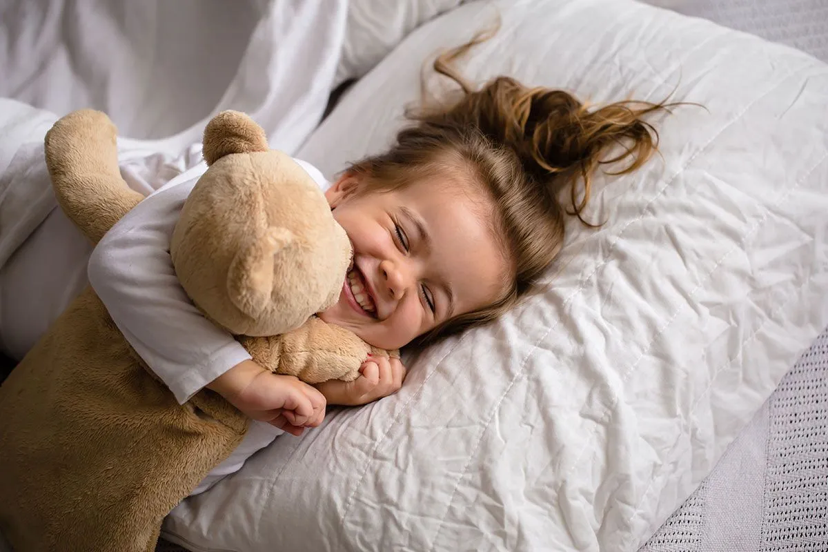 Liten jente som ligger i sengen og smiler og koser med bamsen sin.
