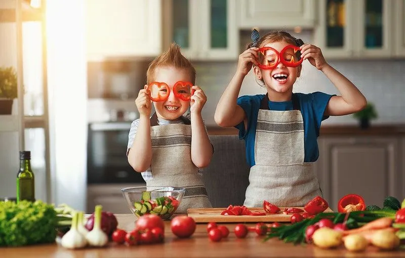 Vaikai virtuvėje daro juokingus veidus su salotomis ir juokiasi. 
