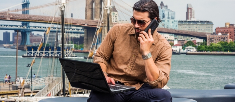 Hombre de negocios estadounidense viajando, trabajando en Nueva York, usando camisa marrón, gafas de sol, sentado en un banco en el puerto