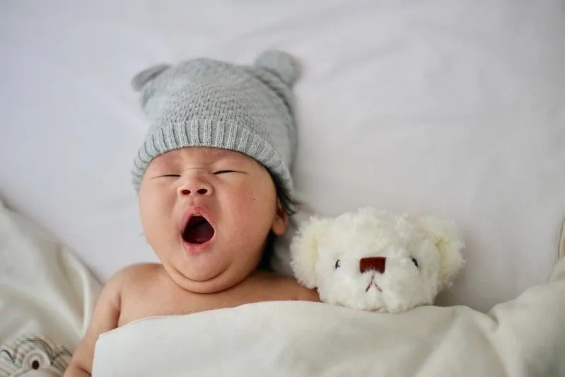 Liten baby i ullhatt i sengen som gjesper ved siden av en bamse.