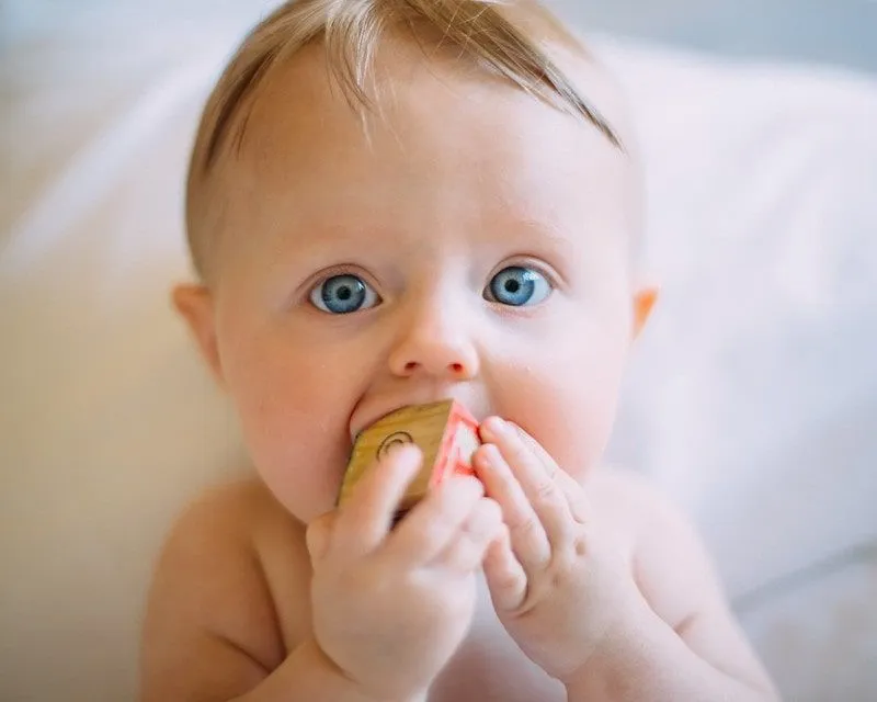 Mėlynaakis kūdikis čiulpia žaislinę kaladėlę.