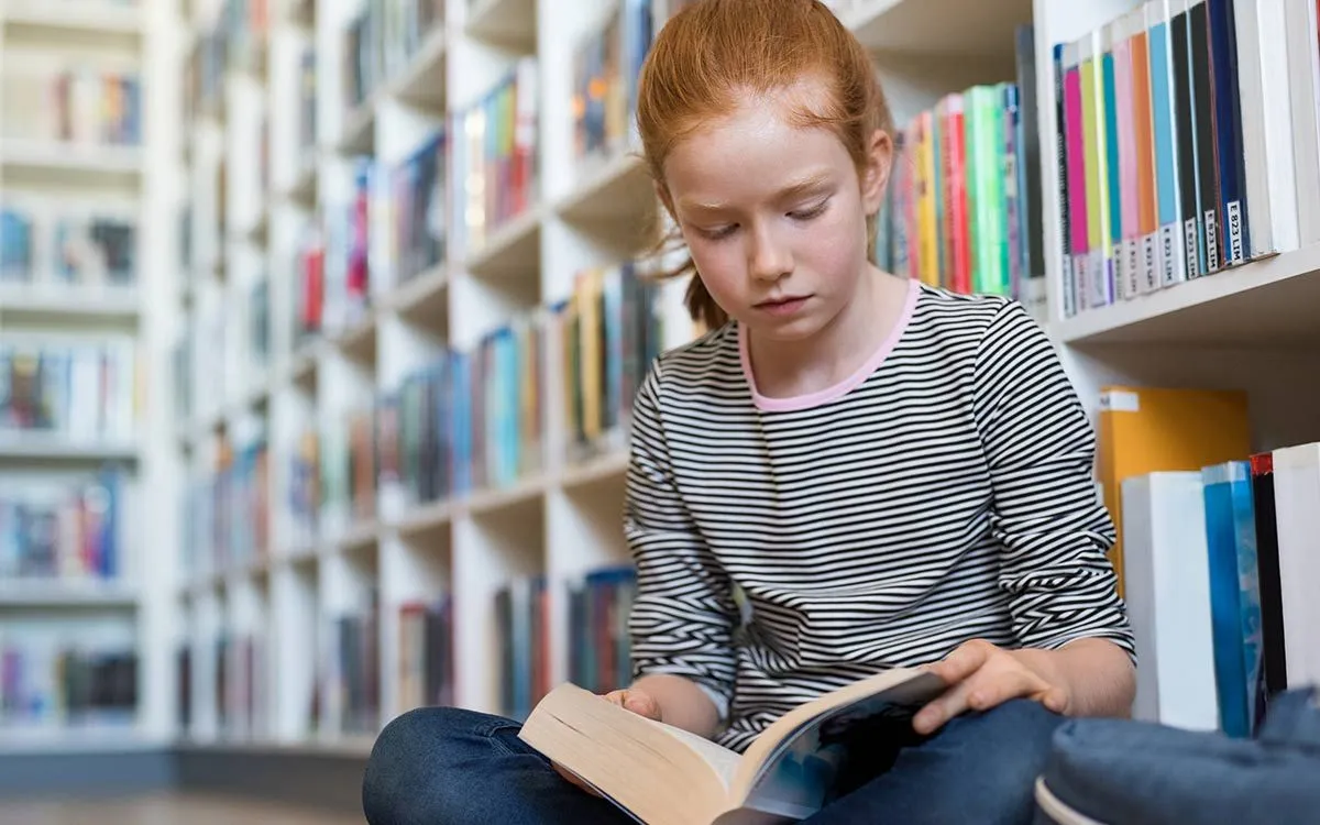 Jauna mergina KS2 sėdėjo ant grindų bibliotekoje ir skaitė knygą, padedančią atpažinti vaizdinę kalbą.r