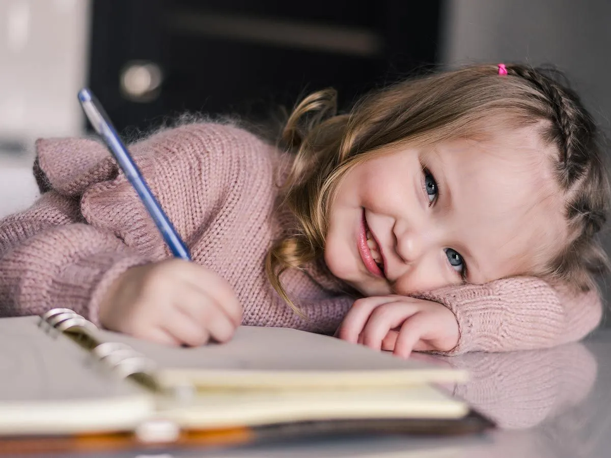 Una niña que escribe en un cuaderno mira hacia arriba y sonríe a la cámara.