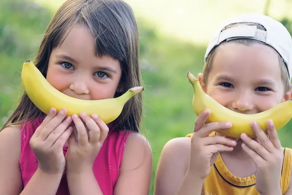 Μικρό κορίτσι και αγόρι στέκονται έξω κρατώντας μια μπανάνα στο πρόσωπό τους ως χαμόγελο.
