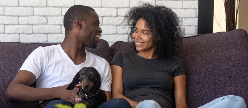 Svarte familieleietakere feirer flytting sittende på sofaen med hund, huseiere som har det gøy å pakke ut i eget hus