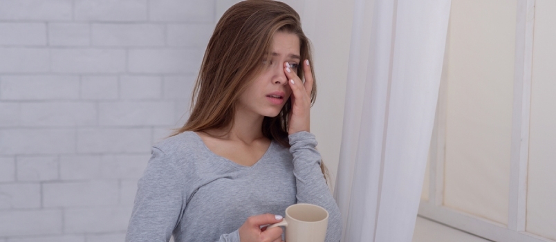 Giovane donna infelice in piedi vicino alla finestra con una tazza di caffè e piangendo, all'interno