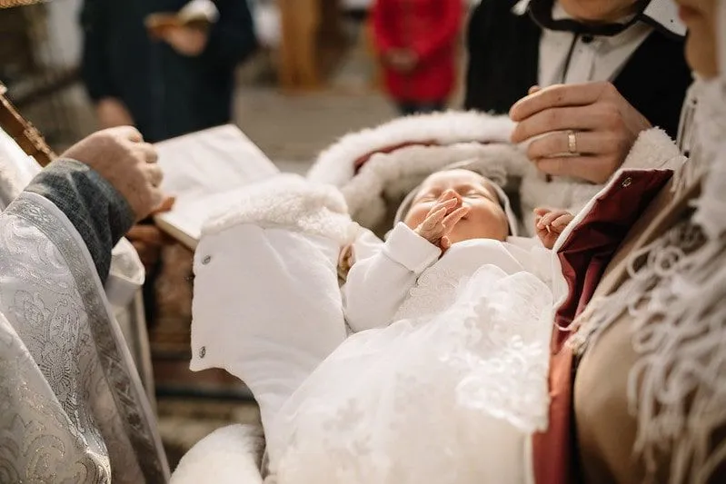 Беба обучена у бело, лежи у наручју родитеља пред свештеником на крштењу.