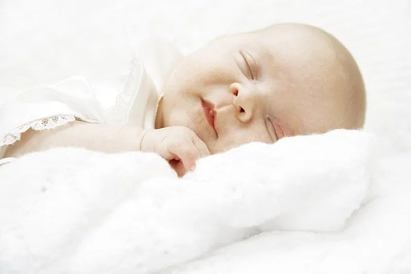 Новорођена беба обучена у бело, спава на белом кревету.