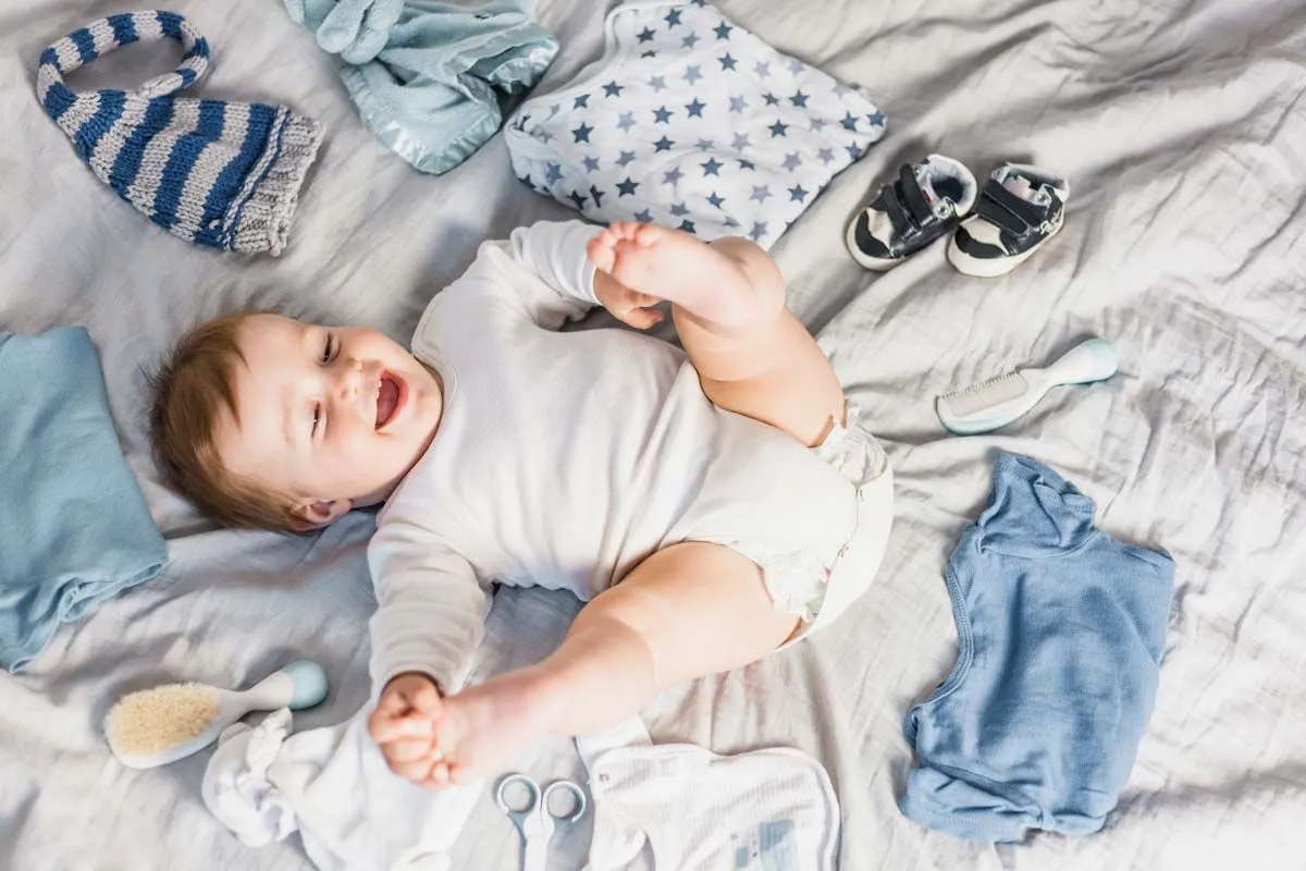 En baby gutt ligger på en seng og ler, han er omgitt av masse blå klær.