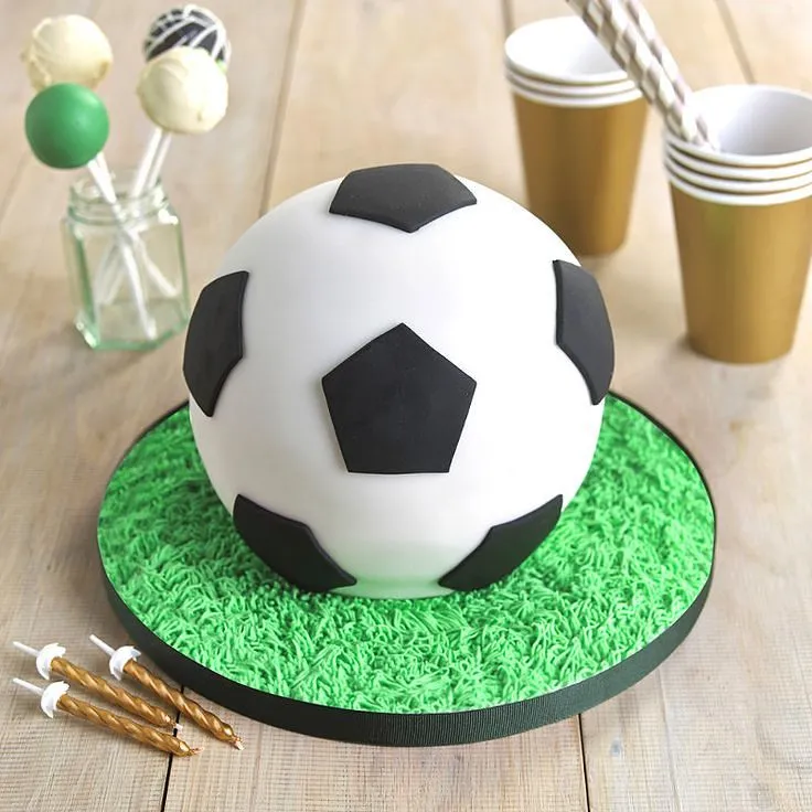 Tårta formad och dekorerad som en fotboll, serverad på en bräda av grön glasyr som liknar gräs.